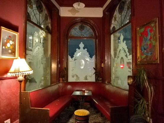 A trapezoidal space, framed by a triptych of windows and furnished with a table and benches in early 1900s style. The prevailing colour is red/brown.