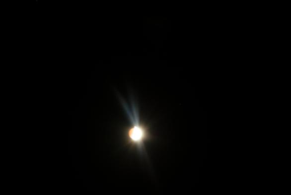 start of the lunar eclipse.
the moon is in the lower middle.
an almost complete yell0w-white ball
a crescent of copper coloured eclipse can be seen on the left of the ball.
lense flare streaming above and bellow the moon can be seen.