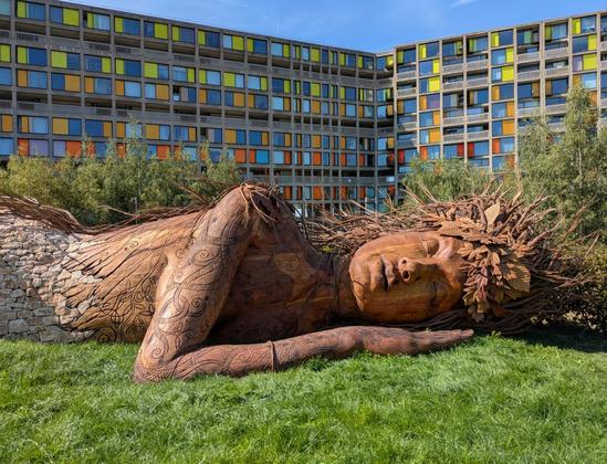 A large wooden statue of a recumbent woman lies on grass in the sunshine. Her unkempt hair  has a headband of carved leaves. Her right arm lies across her body and lifts her head slightly off the ground. Her and arm are carved to suggest intertwined tattoos. Her lower body transforms into a dry stonewall. In the background between the blue sky and a hinterground of green bushes is a complex of highly coloured façades on a brutalist housing complex. The statue is the representation of Gaia from "Gaia Sleeps Amid", Sarah Eberle’s 2026 Award-Winning Garden at the Royal Horticultural Society, Chelsea Flower Show.