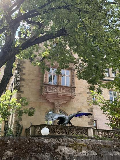 Huge bronze statue of an eagle with its wings spread on the balcony of an old house with a beautiful window over it and a tree giving it shade