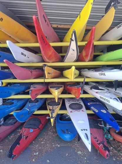 Dans un hangar, des kayaks de toutes les couleurs rangés sur des étagères. Certains ont la pointe qui remontent,  on dirait des tranches de fruits.