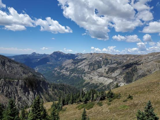 A mountain valley seen from the pass at its head. Nearby the slop is relatively flat, covered in brown grass and green pine trees. Beyond that the ground drops away into a steep cliff as it descends into the valley. The valley has a glacial carved U-shape and its base is covered in trees. The sky above is a brilliant blue with fluffy clouds. On the horizon you see many additional mountains