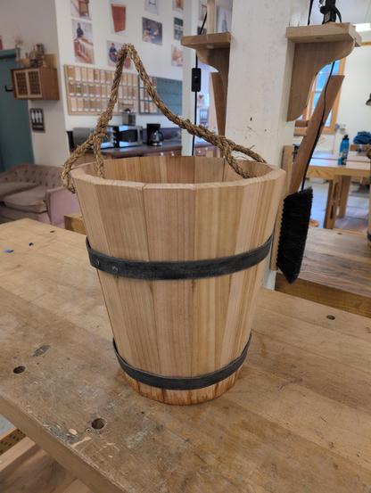 A bucket made out of red cedar wood. There are 16 staves (planks) arranged vertically which taper to be wider at the top, allowing for the bucket to have outward sloping sides. There are two steel bands (hoops) that hold it all together. It has a rope handle that I spliced loops into to attach to the bucket.