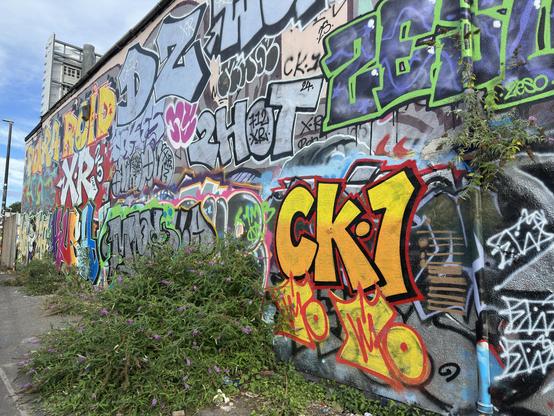 A wall covered in graffiti text, with two Ck 1 tags, one large and  drawn with time and car, the other rapidly sprayed. Both done over a decade after his death