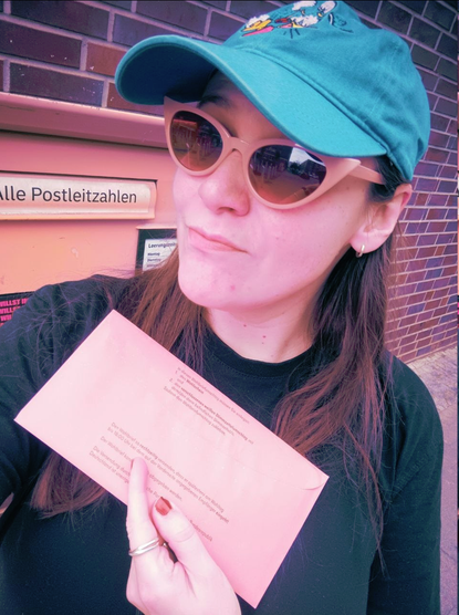 A photo of myself wearing a blue Donald Duck cap, pink sunglasses and a plain black shirt. I have medium brown hair and I'm standing in front of a post box, holding my ballot up in one hand. The photo has a slight cyberpunk filter that makes everything look more pink and blue-ish.