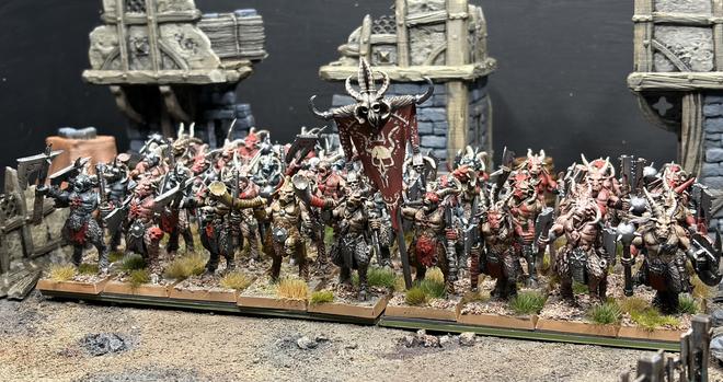 A large group of goat men dressed in minimal armour with all kinds of jagged weapons emerging from a ruined town. Varying colours of fur and flesh, unified by red armour, war paint and a skull bedraggled banner.