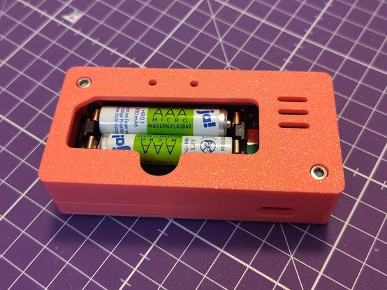 A pink 3D printed case with an opening  through which two NiMH AAA batteries can be put into the battery holder of the charging PCB. 

A half round cutout is supposed to help with reaching the batteries. 

The background is a purple cutting mat.