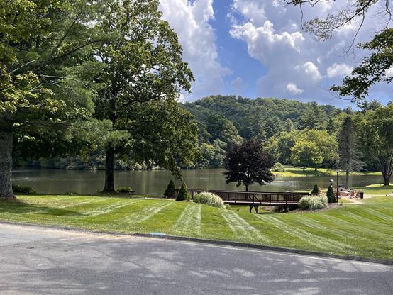 Peaceful scene of a small resort lake surrounded by manicured grass, trees, a  walking bridge with slatted sides, and green shrubbery, with a tree-covered hill in the far background.  Sunlight highlights a grove of trees & grass in the mid-distance on the far side of the lake. The sky is blue with white and gray billowing clouds. The foreground is gray asphalt of a driveway.