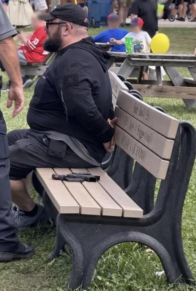 A heavy set man clad in a dark sweatshirt, shorts, and cap is handcuffed and sitting on a bench after apperently attempting to attack candidate Dr. Amy Acton at a campaign event.