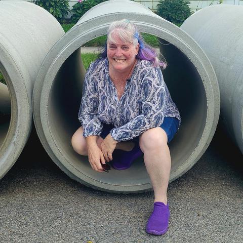 An older white woman with shoulder length grey hair partially dyed blue and purple wearing bright purple shoes, denim shorts and a long sleeve blue and white patterned shirt, sits crouched inside a concrete water tube that's about 3 feet diameter. It's one tube in a row of tubes. I'm smiling broadly and slightly flushed from the heat.
