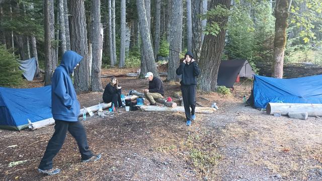 Campers huddle around the stove and bear cans eating. One is standing with her tongue stuck out at the camera.