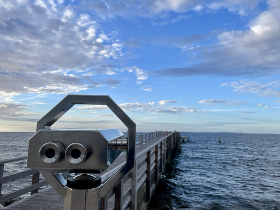 Farbiges Querformat. Himmel mit Wolken, Meer, Steg und fest montiertes Fernglas, das aussieht wie ein Marsianer in einem billig gemachten Science Fiction Film oder einem KI generierten Video zur Erläuterung des Meeresleuchtens.