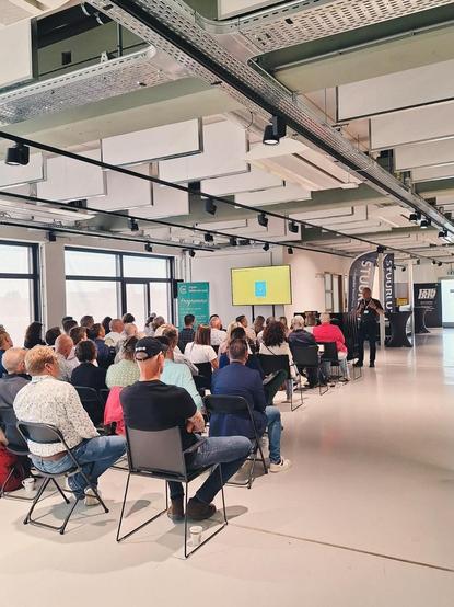 Foto van een zaal waar mensen op een stoel luisteren naar een spreker.