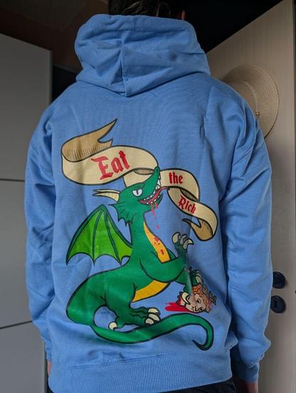 Eine Person steht mit dem Rücken zur Kamera. Sie trägt einen Kapuzenpulli mit einem riesigen Aufdruck:
Übet einem Drachen schwebt ein Banner mit dem Text "Eat the Rich". Dem Drachen tropft Blut aus dem Mund, in der linken Vorderklaue hält er einen abgerissenen Kopf mit Krone.

Der Kapuzenpulli ist noch zerknittert vom Versand. Im Hintergrund ist eine Tür zu erahnen, an der ein Sonnenhut hängt.