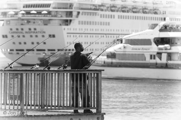 This is a black and white street photo in landscape format of an African-American man fishing with eight rods at the end of a small pier. Beyond is a SeaCat fast ferry underway and a moored cruise ship in the background. Long Beach, California (2015).
Extending from the lower left corner to the middle of the photo and rising upwards to the half way up level are the weathered metal railings of a small pier with the wooden flooring just visible at the base of the image. Standing in the right corner of the pier (and in the centre of the photo) is our mid-twenties (very early thirties) African-American man. He is viewed obliquely with his back and right side in view, he is filling half the height of the photo. The camera is at the same level as the man and positioned four to five metres away. He has short neat hair, a short trimmed beard, is wearing a dark, long-sleeved sweat shirt and jeans. In front of him and attached to the railings are seven fishing rods, their thin mono filament fishing lines are stretched outwards into the water ahead and below. In his left hand is the eighth fishing rod, the line, again, out over the water and he is holding the reel in his right hand. In the lower right corner, the water stretches upwards to the one-third up level. Extending from the right edge of the phot to the centre is the white SeaCat fast ferry underway from right to left. The ferry is slightly blurred out of focus but the two decks are visible with the wheelhouse on top. The vessel's name 'Catalina Express' can just be made out. The background is dominated by the stern and starboard side of a large white moored cruise ship. Again this ship is slightly blurred out of focus but the name 'Carnival Inspiration' can be read high above her stern.