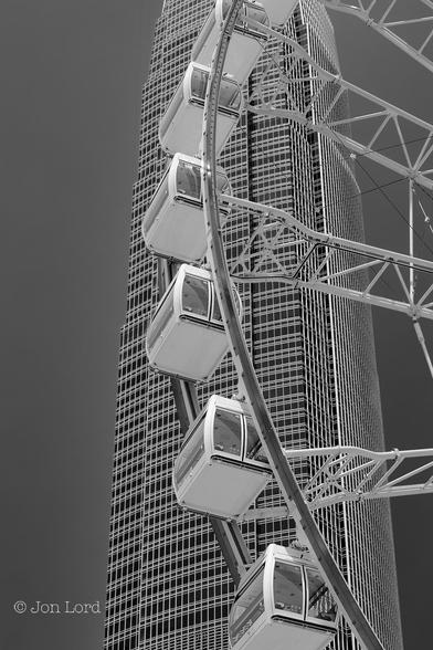 This is a black and white abstract architectural photo in portrait format of a portion of a Ferris Wheel in the foreground and a modern high-rise office block in the background. Central, Hong Kong (2015).
In the foreground is a small section of the Hong Kong Observation Wheel. In view are six white cabins, two large rings that circle the wheel and four lattice spokes that attach the outer rings of the wheel to the central hub. Each cabin is a horizontal rectangular box with large windows and attached to the two rings by a gimbal on the roof so that the cabins stays upright at all times. Behind is a the 2IFC, a 412m high-rise office block. The tall and narrow building has a nearly square footprint at the base but with slightly curved convex glass fronted sides. While the building has 88 floors above the ground the numbers 14 and 24 are missing since in Cantonese culture these numbers are considered inauspicious. In the background is a clear and cloudless sky rendered a dark grey.