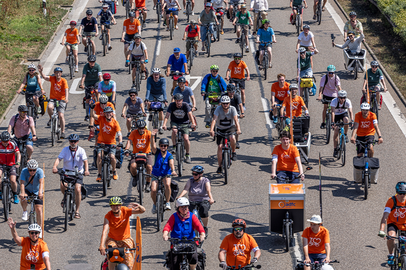 Viele Radfahrende fahren bei der Sternfahrt in Stuttgart auf einem für Autos gesperrten Autobahnabschnitt. Viele tragen orangefarbene T-Shirts des ADFC, im Vordergrund ist ein Lastenrad mit ADFC-Logo zu sehen. Mehrere Personen winken in die Kamera.