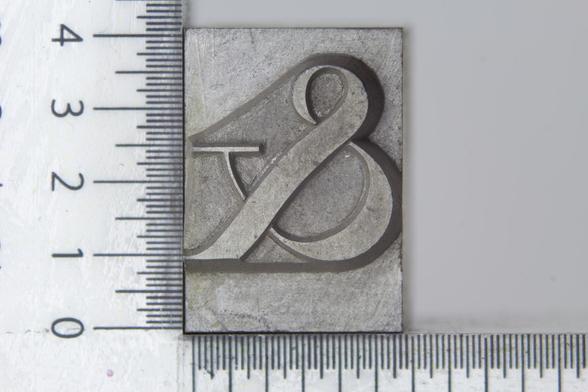 Close-up photograph showing a metal-type ampersand sort, with ruler units for measurement on the left and bottom sides. The ampersand is from the Tiemann typeface in the 48 pt. size.