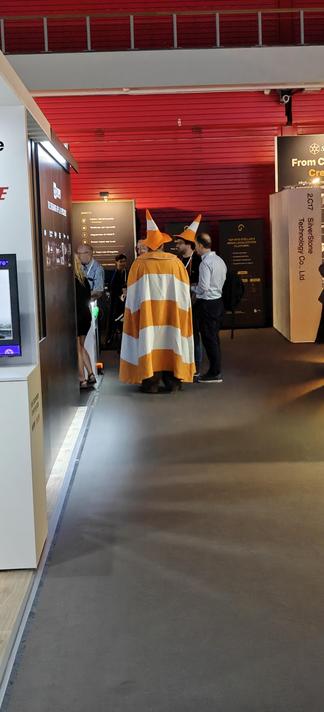 Man seen from behind wearing a traffic cone on his head, and with a cape with broad white and orange horizontal stripes, resembling the VLC media player icon