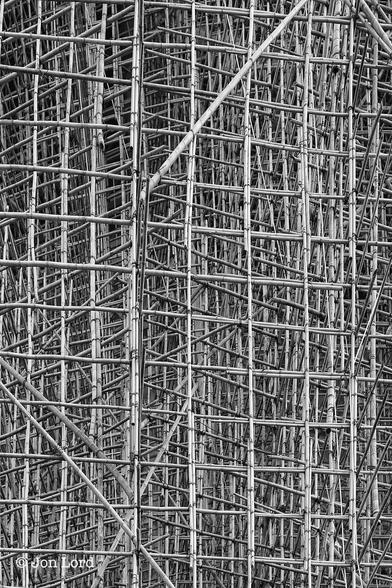 This is an abstract black and white photo in portrait format of an enormous framework of bamboo scaffolding surrounding the west side of the Hong Kong Convention Centre in Wan Chai (2014).
Filling the entire photo is this vast frame of multiple poles of bamboo attached to each other with lengths of plastic binding straps. The camera is set back from the structure by no more than ten metres and is angled directly at the frame. The base, sides and top of the scaffold are out of view outside the edges of the photo. The bamboo is fashioned into squares of about one metre in length and height. Each pole overlaps the next pole by about two metres with that area secured using numerous binding straps similar or identical to those used to wrap a large package. In view are thirteen horizontal lengths and eight vertical poles. Behind those lengths at the front is a vast matrix of further poles extending towards the building. Most of the bamboo is either vertical or horizontal, or stretching inwards towards the building forming cubes out of the bamboo, though there are some diagonal lengths for stability. It is hard or impossible to say how many frames there are between the first and the building but it must be at least five.