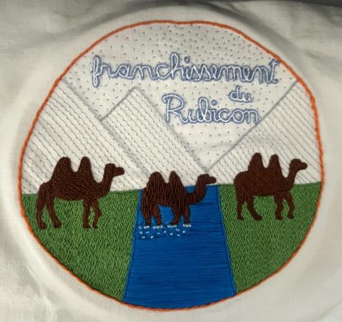 three camels, one on each side of a river and one currently crossing it. above it in the sky is written “franchissement du Rubicon”. The grassy river banks, the river, and the camels are very densely stitched with bold colors but the distant mountains and sky are very sparsely stitched with light colors and there is a lot of white linen background visible above the horizon.