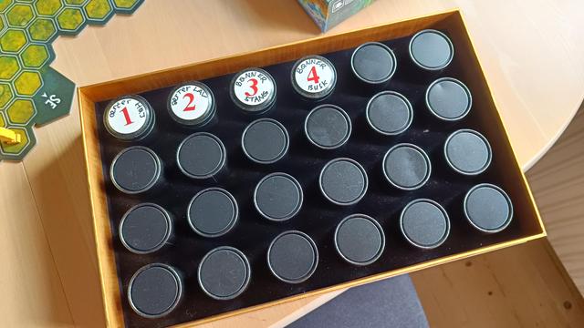 Pic of the start of my seed collection, shown on a sunny table next to a board game with game play in progress (Aspens)

24 small metal tins arranged in neat rows of 6x4 rest snug inside of last year's tea advent calendar box

The tins are black aluminium cylinders, stored bottoms up except for the first 4, which are flipped up and annotated with the Danish names for what's inside

Left to right these read
1 - lav ærter (low growing peas)
2 - høj ærter (tall/runner peas)
3 - stang bønner (runner beans)
4 - busk bønner (bush beans)