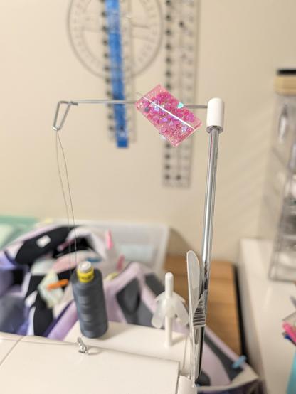 An antenna that holds thread up with spaces for two spools, attached to a sewing machine. A bright pink glittery magnet with a hand sewing needle is stuck to the empty guide.