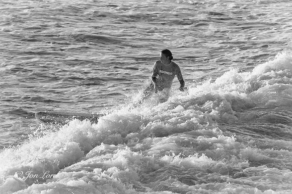 This is a black and white sports photo in landscape format of a young woman surfing at sunrise. Hermosa Beach, California (2013).
Stretching diagonally across the photo from the lower left corner towards the upper right is the foaming surf of a breaking wave that's moving from right to left. In the centre of the image is our teen surfer, surfing across the wave from right to left. She is viewed from about five metres away and two metres above and with her filling about half the height of the photo. Her body is facing the camera while her head is angled ahead, inline with the surfboard. She has dark hair that is tied behind her head, is wearing a light toned t-shirt with 'Redondo' printed on the front and covering her wetsuit. Below her waist is obscured by the breaking wave. Just visible ahead of the wave is the forward portion of her white surfboard that has two parallel black stripes inset from the edges. The sea ahead of the breaking wave is heavily disturbed with multiple small wavelets.