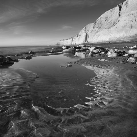 A black and white photograph of a pool of water on the beach at Torrey Pines, San Diego, California. Beach cliffs rise tall in the distance.