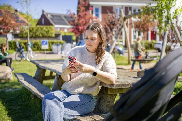 Een jonge vrouw zit aan een picknick-tafel. Ze heeft een smartphone in haar hand. Op de achtergrond een speeltuintje en huizen.