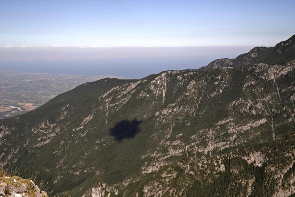 A mostly forested ridge with some rocky outcrops and the sea behind. Also a distinct round cloud shadow.