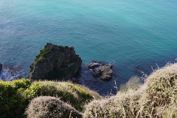 Most of the picture is of water that is a deep azure colour along the rocky shore. There are some flat rocks and a small spire in the water. At the bottom of the picture there are some yellow and green plants.