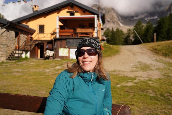A person standing in front of a refuge in the Alps. I write "refuge" but it's a pretty sturdy and not basic building, basically a modest hotel. There is a tiny bit of blue sky but it's mostly clouds behind the refuge, and some Alps are peeking through.