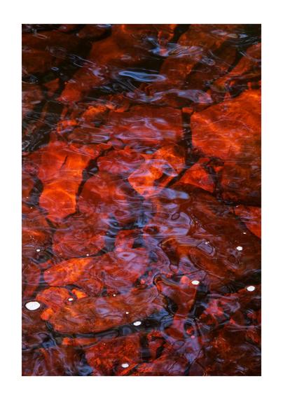 A portrait oriented photo looking down on a rocky riverbed, with feint water ripples, swirls, and reflections visible on the surface of the water, which is a deep red-orange colour. There are also small clusters of white bubbles on the surface.