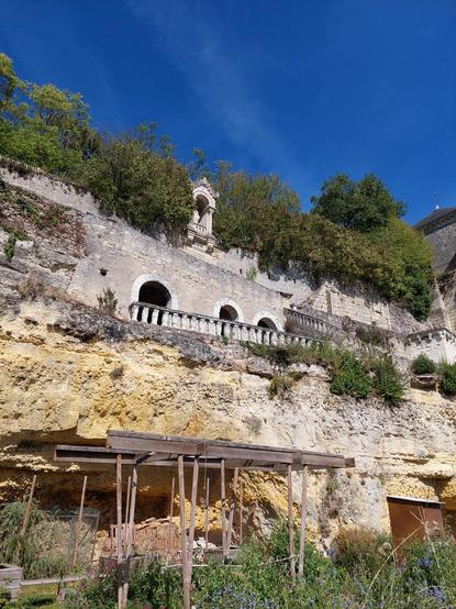 Un coteau creusé étayé et des murs, des escaliers, des ouvertures en ogives surplombés de végétation sous un ciel bleu éclatant