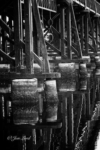 This is a dark and grungy black and white photo in portrait format of an old wood and metal bridge spanning Cockle Bay. Sydney NSW (2013).
The lower half of the photo is filled with still, jet black harbour water with the occasional ripple breaking the surface and numerous, confused silver reflections of the bridge above. The camera is located a metre or two above the water, two metres from the front of the bridge and angled along its length. Sunk into the harbour are multiple concrete pillars with about one metre exposed above the water's surface. While the tops of the pillars are white, all that is below is dark and covered with a thick layer of marine growth from the rising and falling tides. The pillars support hundreds of long, square and black posts. Some are vertical, some diagonal and some angled slightly off the vertical. All are joined with countless long black metal bolts. The black wooden posts extend upwards around three metres and then supports horizontal metal lattice frames that form the base of the bridge deck. Beneath one span hangs a maritime symbol made from one black ball, a black triangle and a further black ball, perhaps suggesting reduced vertical clearance?