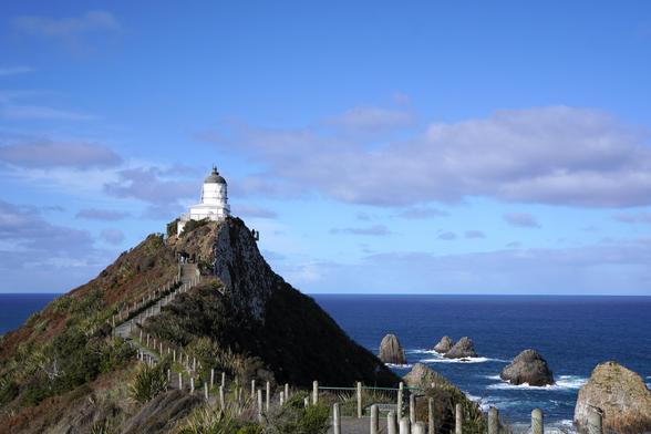 Change of pace. A white lighthouse on a hill on a peninsula. The lighthouse has a grey/copper green roof. Around the peninsula there is dark blue water with some smaller rocks sticking out.