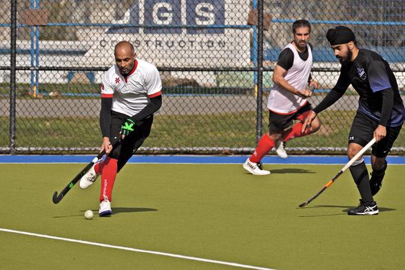 A person wearing a white shirt, black shorts, and red socks is controlling the ball with his stick. An opponent with a black shirt looks on. There's another person in the background with the same white shirt.