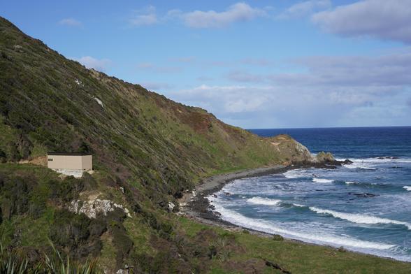A boxy beige hoiho hide looks over a bay with a couple of waves and, if the time is right, some hoiho/yellow-eyed penguins.