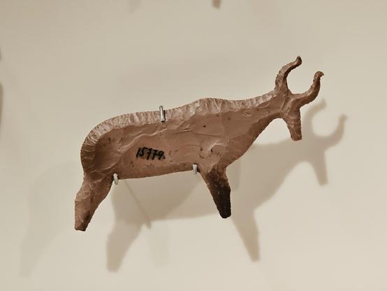 A flint figurine shaped like hartebeest. The objcet is brown and has a rough, chipped texture. It is mounted on a display with metal supports, casting a shadow that emphasizes its form.