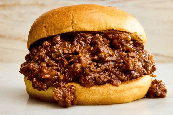 A soft sandwich bun is stuffed with a heaping mound of saucy ground meat filling that spills out over the sides. The background is a light, neutral surface.