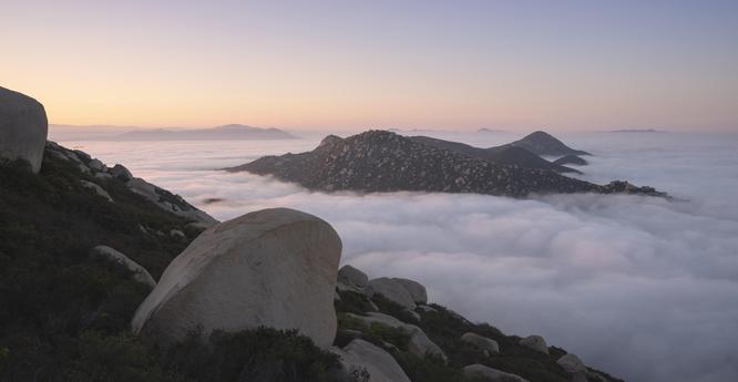 A color photograph of a sunrise watched from the steep slopes of Mount Woodson in East County San Diego, California. Marine layer clouds stayed low with Iron Mountain popping up in the distance.