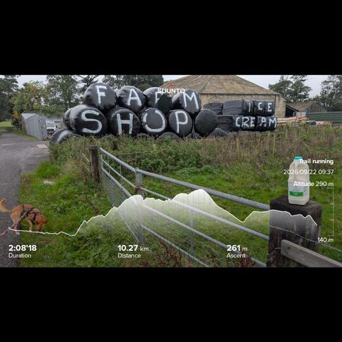 Photo of a farm with big black plastic silages bails stacked up with Farm Shop written on them. In the foreground in a fence post I've put the 2litres of milk and Penny is busy with sniffing the grass for either little critters or rabbit poop. My running stats imprinted on the top