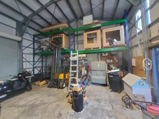 Wide angle view of the tidier end of my workshop. Racking up to the ceiling. Forklift under it on left, roller door further to left. Other stuff kicking about including batteries, bins, bits and bobs.