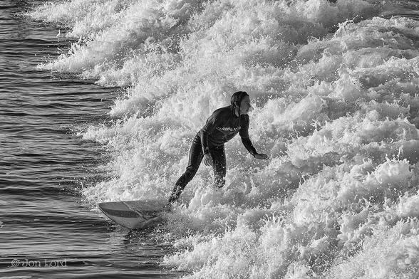 This is a black and white water sports photo in landscape format of a female teen surfing shortly after sunrise. Hermosa Beach, California (2013).
All of the right side of the image, extending from the centre of the base of the photo, diagonally upwards to the upper left corner: is brimming with a maelstrom of foaming brilliant white surf from a Pacific wave breaking from right to left. The remains of the image, to the left, is filled with jet black sea water with horizontal ripples disturbing the surface. In the centre of the picture is our female teen. She is viewed from a few metres above, three metres away and with her filling half the height of the photo. She is standing on her surfboard, body facing the camera, her head angled back as she looks at the breaking wave behind her, perhaps with an anxious expression on her face. She has long, dark and wet hair, is wearing a dark t-shirt with the word 'Redondo' in large white lettering and a black wetsuit underneath. She is surfing from right to left and slightly towards the camera.