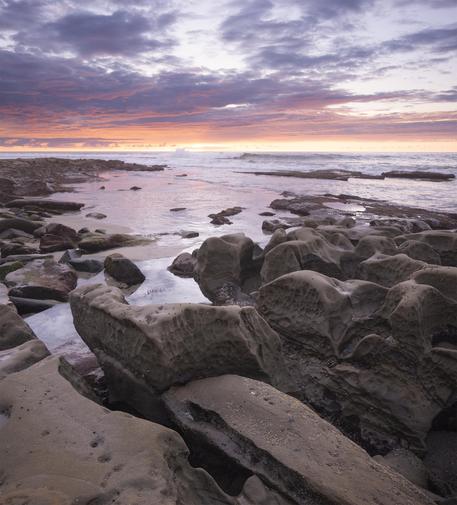 A color photograph of rock formations along the coast of La Jolla, San Diego, California. Clouds lit up in warm shades at sunset.