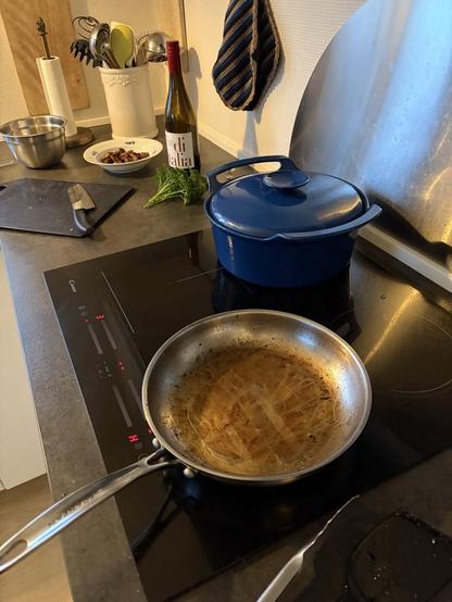 Steel frying pan with golden-brown residue in the foreground, then a blue enamel cast-iron pot, a small bunch of parsley, a wine bottle (whose erstwhile contents are mainly in the blue pot and the cook) and a small dish of fried mushrooms.