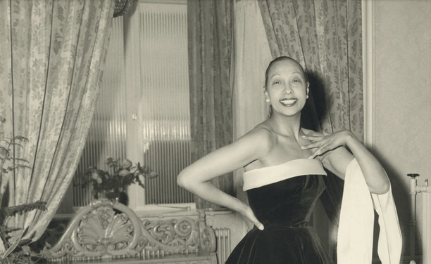 A smiling woman poses elegantly in a strapless dark dress with a light trim, one hand on her hip and the other near her shoulder. She stands in a room with patterned curtains, a window, and ornate furniture behind her.