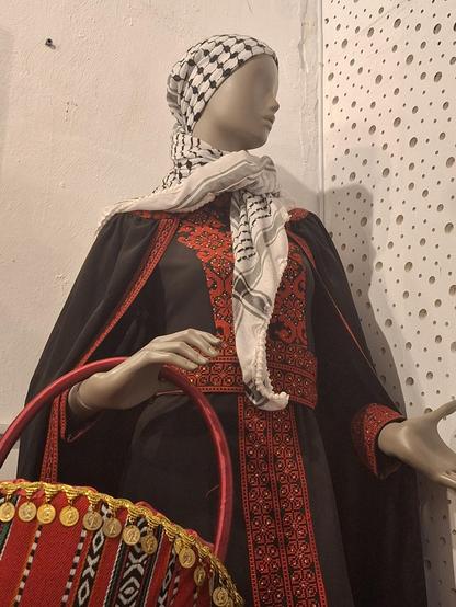 Fotografía de un maniquí con un vestido negro y rojo clásico palestino y una kufiyah en la cabeza. Lleva además una cesta en el brazo, también en negro, rojo, verde y blanco.