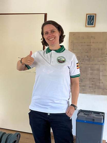 I am standing in a room and proudly wearing a white polo shirt with green color and sleeves, with the Eco Youths Network logo on the front, and a Ugandan flag on the left arm.
I smile happily and give a thumbs up.